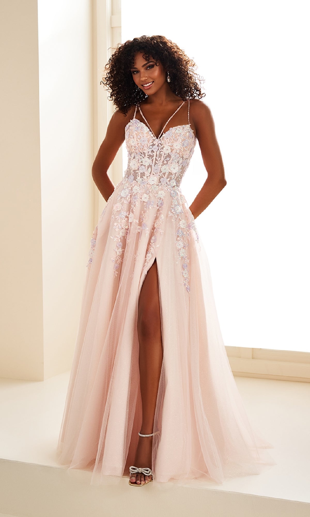 A woman wearing the Ellie Wilde EW36079 long prom dress in light pink, featuring a corset bodice, floral lace details, spaghetti straps, and a thigh-high slit, stands indoors smiling.